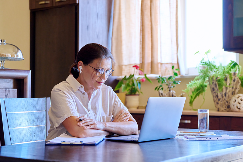 Serious mature woman in headphones talking on video using laptop. Middle aged business female working at home at the table, communication consultation. Psychologist on online therapy with patient